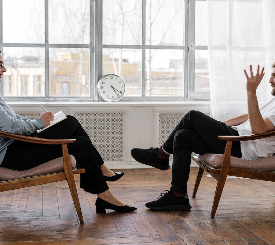 Person in Black Pants and Black Shoes Sitting on Brown Wooden Chair